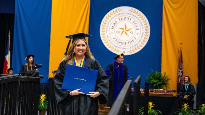 Student smiles with diploma in hand and graduation cap on her head. The university's seal and the graduation stage are behind her.